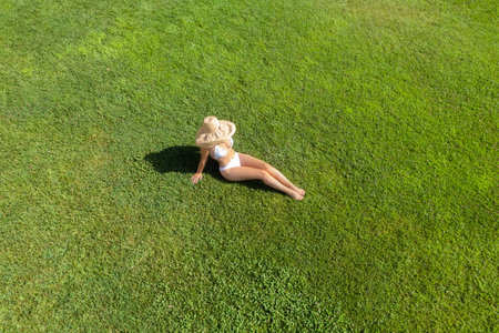 Woman in a white bikini and hat, sitting on the green grass sunbathes at summer day. Top view, drone, aerial viewの写真素材