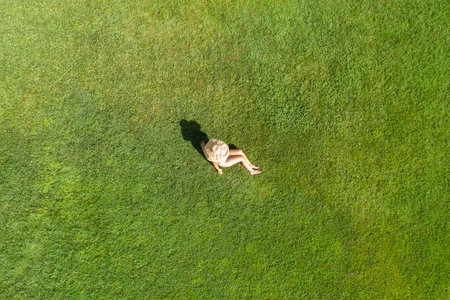 Woman in a white bikini and hat, sitting on the green grass sunbathes at summer day. Top view, drone, aerial viewの写真素材