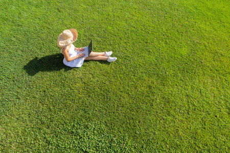 Woman in a white dress, hat with laptop, sitting on the green grass at summer day. Top view, drone, aerial viewの写真素材