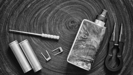 black and white, monochrome shot of coil jig, 18650 battery, fused clapton coil, box mods with rebuildable dripping atomizer, black ceramic tweezer on wood texture background, focus on an attyの写真素材