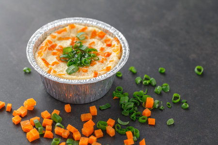 steamed egg with diced carrot and chopped spring onion in aluminium foil cup on dark texture...