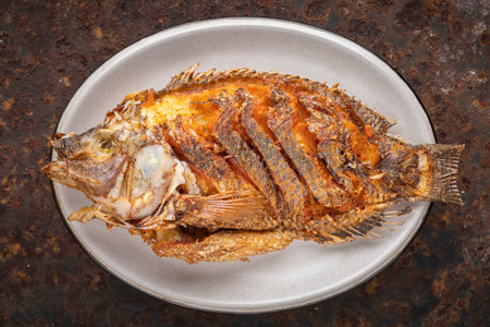 tasty large fried nile tilapia fish in oval ceramic plate on rusty texture background, top view