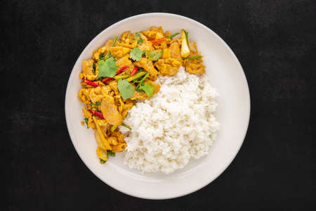 Stir Fried Chicken with Curry Powder beside Steamed Rice in simply white ceramic plate on dark tone texture background, top view, Gai Pad Pong Karee, Thai foodの写真素材