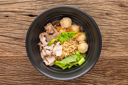instant noodles soup with beef, meatball, pork liver, water spinach, morning glory, celery, fried garlic and herb in black ceramic bowl on rustic wood texture background, top viewの写真素材