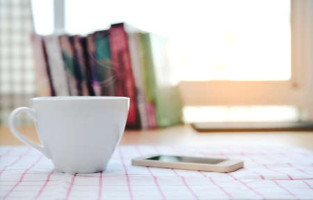 A cup of coffee and a smartphone is on the tablecloth and the book is blurred as the background.の写真素材