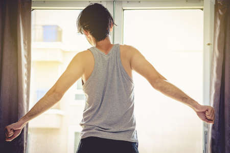 An Asian man opens the curtains and watches the morning light after waking up.の写真素材