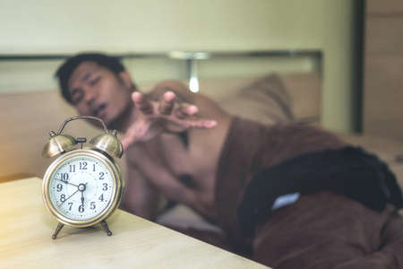 A man lying under a brown blanket is going off the alarm clock with drowsiness.の写真素材