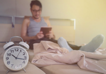 An Asian boy spends playing a tablet on his holiday bed.Focus on the alarm clock