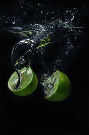 Lime green fruit that is falling and has water spreading with a black background.の写真素材
