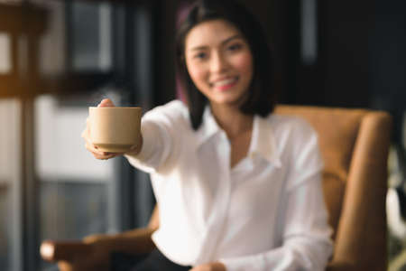 An Asian woman holds a cup of coffee and gives it to invite to drink together.Focus on the coffee cup.の写真素材