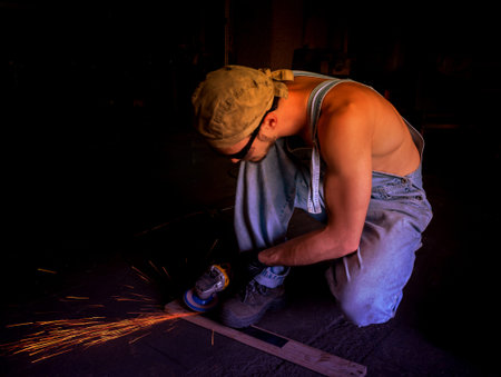 Men at work grinding steel with a grinder in a metal fabrication factory and throwing off bright orange sparks around him while he works.のeditorial素材