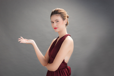 Beautiful woman wearing red dress and showing product on hand in studio gray background. Action for presenting your productの写真素材