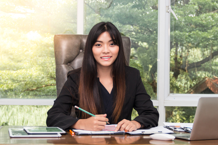 Beautiful young asian businesswoman sitting and working at the table in office and looking with smile at cameraの写真素材