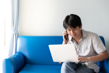 Handsome Asian man using laptop for works and online activities while talking on mobile phone and sitting on blue sofa at the morning timeの写真素材
