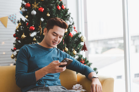 A single man smiling while looking his phone in his living room decorated with Christmas treesの写真素材