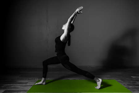 Black white photo of Young beautiful woman brunette in black clothes practice yoga virabhadrasana in dark studio green mat Sport meditationの写真素材