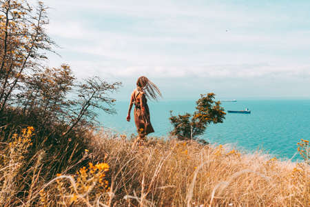 Beautiful slender tattooed woman with blonde dreadlocks in a dress rejoices on a hill. Sea View Travel and Vacation Beautiful landscapeの写真素材