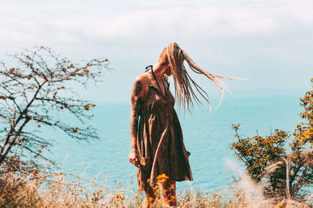 Beautiful slender tattooed woman with blonde dreadlocks in a dress rejoices on a hill. Sea View Travel and Vacation Beautiful landscapeの写真素材