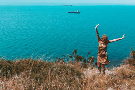 Beautiful slender tattooed woman with blonde dreadlocks in a dress rejoices on a hill. Sea View Travel and Vacation Beautiful landscapeの写真素材