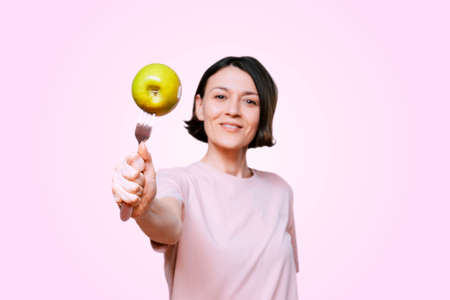 Young Woman holds in hand a fork with green apple on a light pink background. Healthy organic food for dieting woman conceptの写真素材