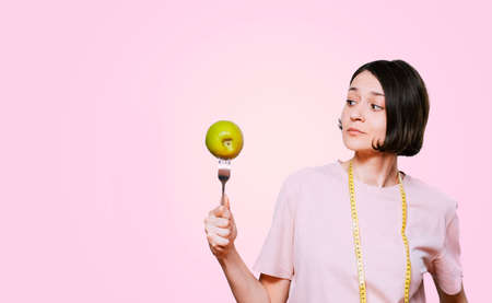 Woman holds in hand a fork with green apple and tape measure on a light pink background. Healthy organic food for dieting woman conceptの写真素材