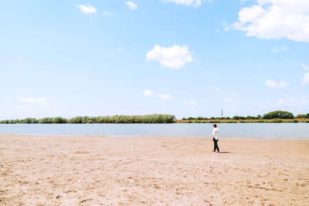 A melancholy girl in a white shirt walks alone by the river Bank. Small figure walks along the sandの写真素材