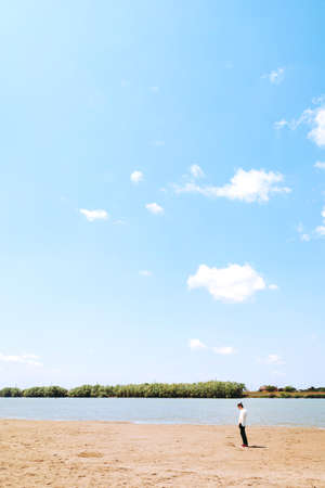 A melancholy girl in a white shirt walks alone by the river Bank. Small figure walks along the sandの写真素材