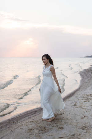 Beautiful young brunette woman in white long dress at the sea shoreの写真素材