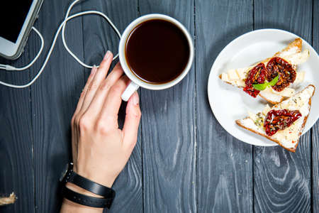 Beautiful background with coffee Cup in hand, toast with sun-dried tomatoes, a mobile phone with headphones on wooden table. The concept of a Breakfast of business peopleの写真素材