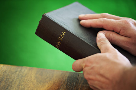 Bible with mans hand resting on the cover whilst holding in front of a green wall
