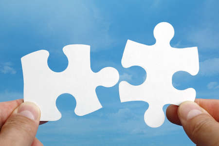 Holding two jigsaw pieces of a blank puzzle trying to fit together against blue sky backgroundの写真素材