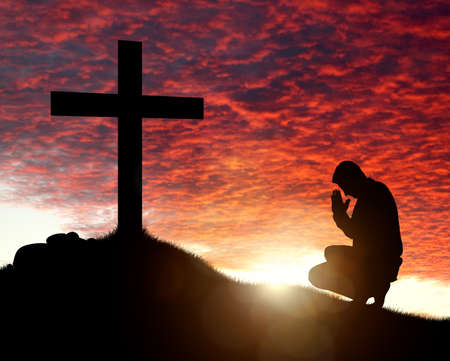 Silhouette of man praying to a cross with heavenly cloudscape sunset concept for religion, worship, love and spirituality