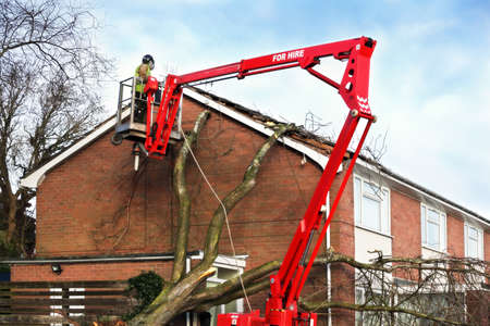 Tree surgeon working up cherry picker repairing storm damaged roof after an uprooted tree fell on top of a residential houseの写真素材