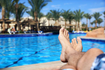 Sunbathing by the hotel tourist resort swimming pool, mans legs lying down on a sunlounger looking over the waterの写真素材