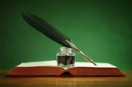 Quill pen and inkwell resting on an old book with green background concept for literature, writing,...