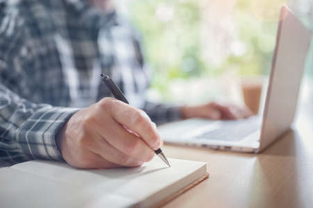 Businessman writing at his desk in office with a laptop computerの写真素材