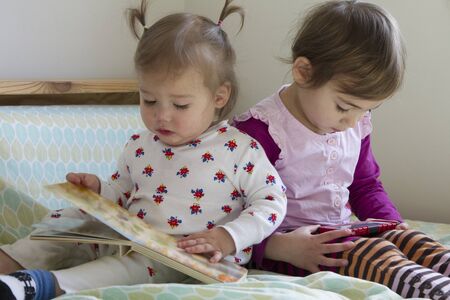 Sisters: 4-year-old girl watching cell phone and 1-year-old girl reading bookの写真素材