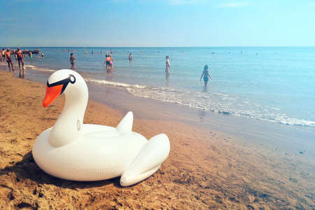 swan toy on the beach, unrecognizable people on the background enjoying summer holidays (inflatable white swan on foreground)の写真素材
