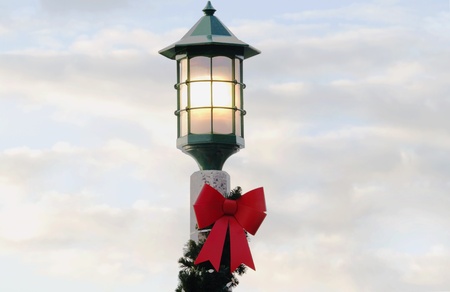 Street lamp decorated with pine boughs and red ribbon is lit against a soft blue and white cloud filled sunriseの写真素材