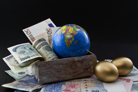 Multiple world currencies including yen, dollar, and euro in a marble box next to two, gold nest eggsの写真素材