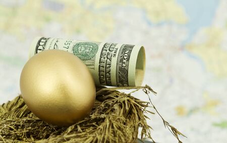 Dollar currency, rolled, sits in gold nest with golden nest egg in front of a map background.  Selective focus on egg and currency.  Map background unfocused through shallow depth of field.  の写真素材