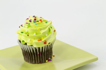 Chocolate cupcake with green frosting and multicolored sprinkles sits on square, green plate;の写真素材