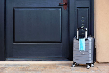 Gray suitcase with  mask placed by black front door reflect necessary pandemic travel preparations