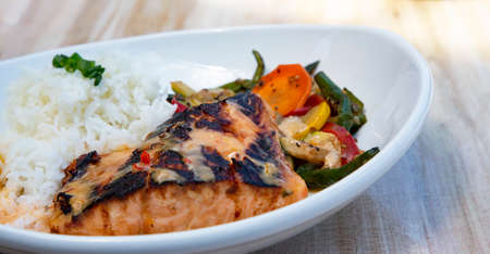 Charred miso marinated salmon with jasmine rice and fresh healthy vegetables on white platter placed on light wood backgroundの写真素材