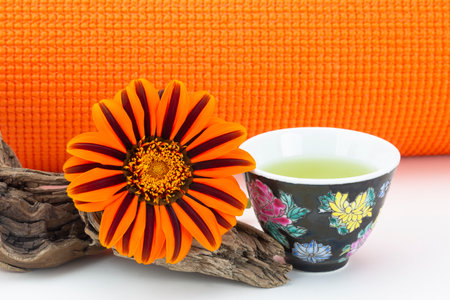 Exercise, hydration, and nature in healthy balance seen in orange yoga mat, natural orange petals of daisy flower, and simple cup of green teaの写真素材