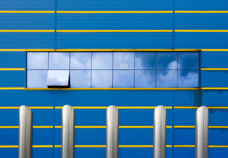 modern blue facade with horizontal lines in yellow. In the middle of the facade a large window reflecting the sky in similar tones to the wall. One of the windows half-open. Ahead, and until rather less half of the vertical,  shining steel columns.の写真素材