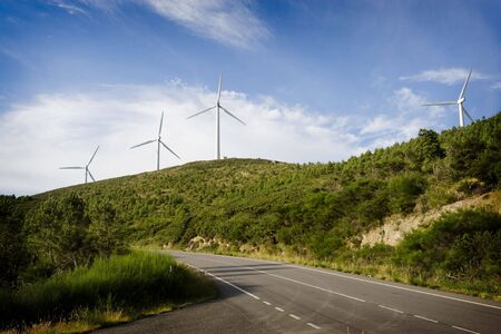 Aeolian generators of electricity on a hill. In first plane a road marks the diagonal of the frame. The sky, to the bottom, is blue intense with clouds. Horizontal frameの写真素材