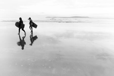 Two bodyboarders walking along the beach with their tables and being reflected in the low tide in one day with mist. Black and witeの写真素材