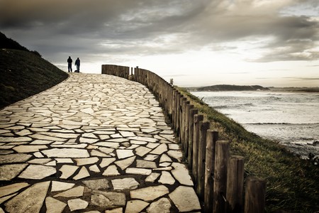 Couple and their dog in silhouette walking along a paved way to shore of the sea. The cloudy day and the hot dominant color they give a romantic aspect to the scene.の写真素材