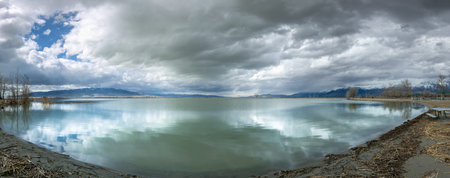 Dramatic cloudscape over calm and reflective Utah Lake with snowcapped mountains in the distance,...